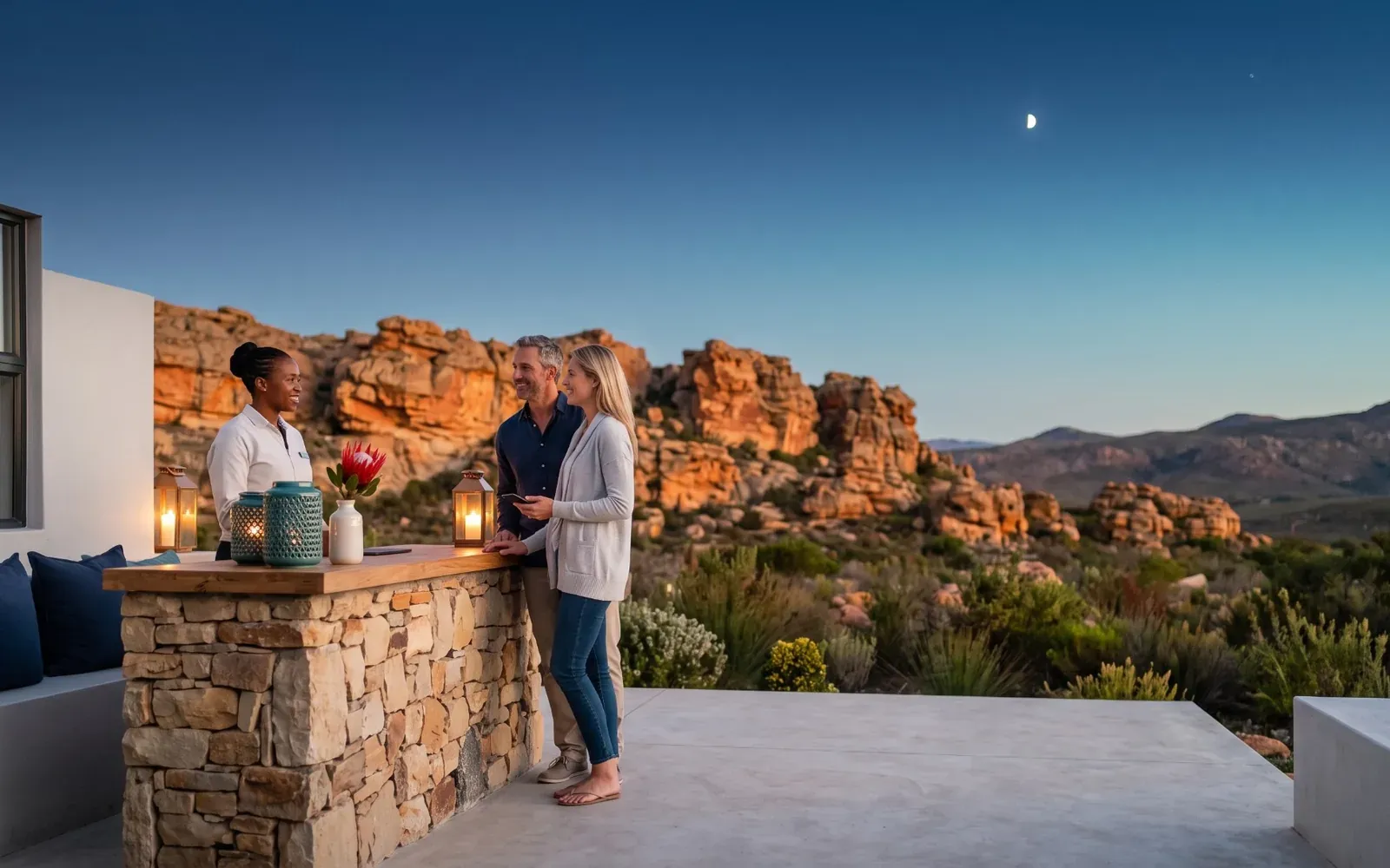 A Cederberg hotel terrace at dusk: a host welcomes a couple at a lantern-lit stone desk, a crescent moon and one bright star above the glowing sandstone rocks